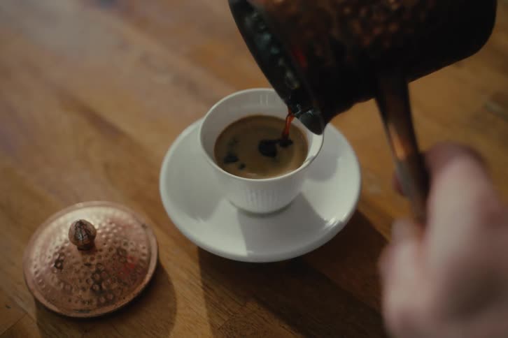 A hand pours turkish coffee from a longhandled pot or cezve