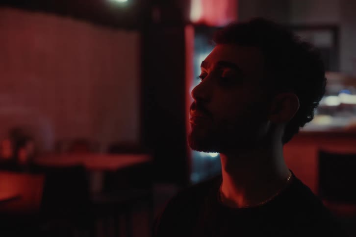 Close up of the profile of a young man standing in a dimly lit room with soft red lighting