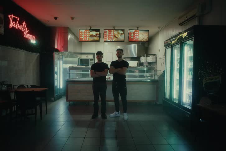 Young people stand around a dimly lit fastfood restaurant with neon signs and large refrigerated beverage coolers