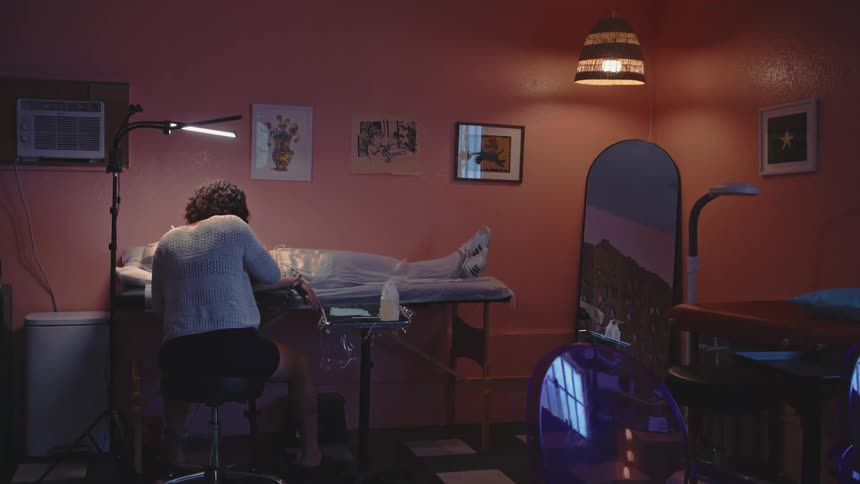 A person is sitting on a stool tattooing a person who is lying on a bed in a tattoo studio