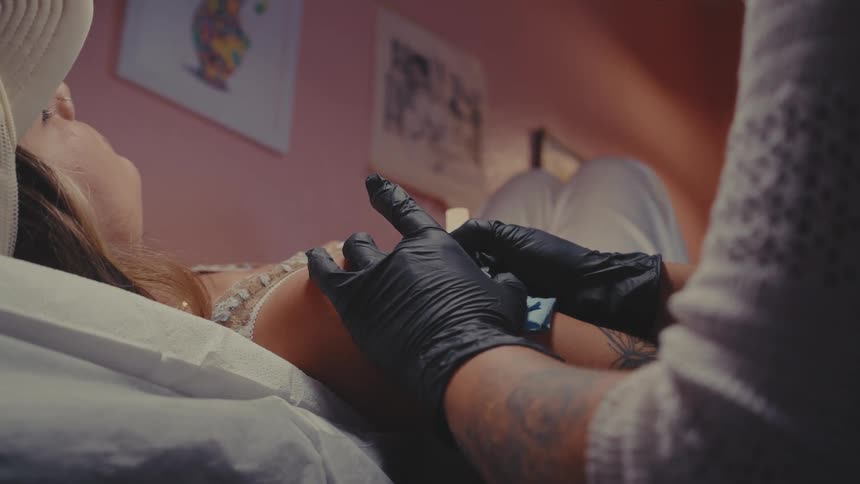 A person receiving a tattoo on their arm from a tattoo artist wearing black gloves