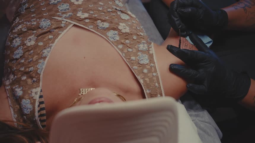 A person wearing black clothes is getting a tattoo on the right arm of a person who is lying down wearing a cap