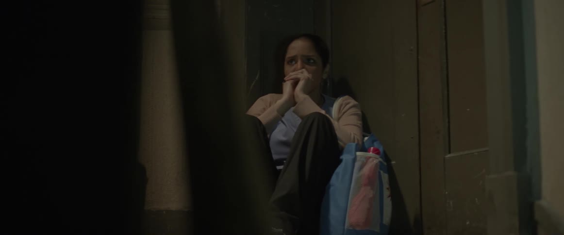 A woman sits in a corner of a room with her hands pulled to her face looking pensive and anxious