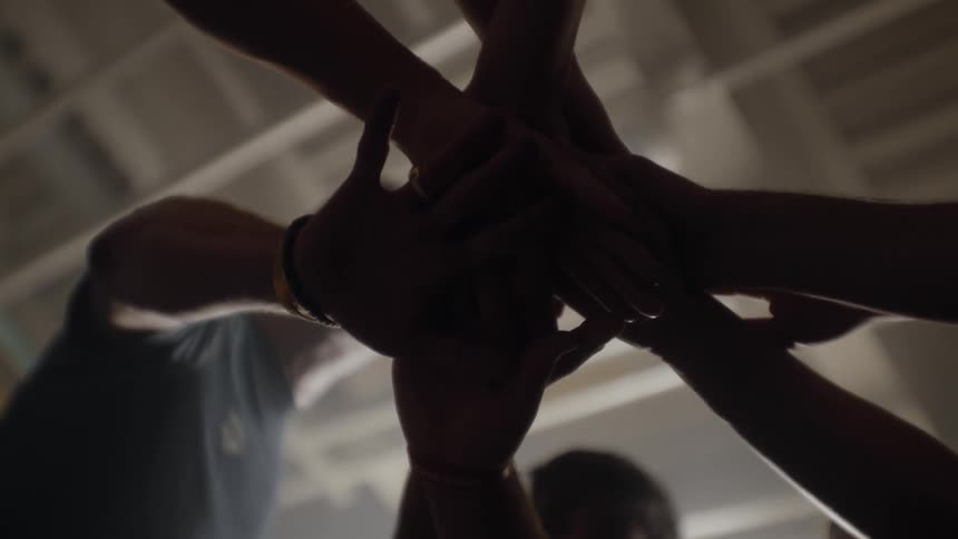 Three people stand in a circle and place their hands in the middle of the circle on top of one another and then stick their arms in the air