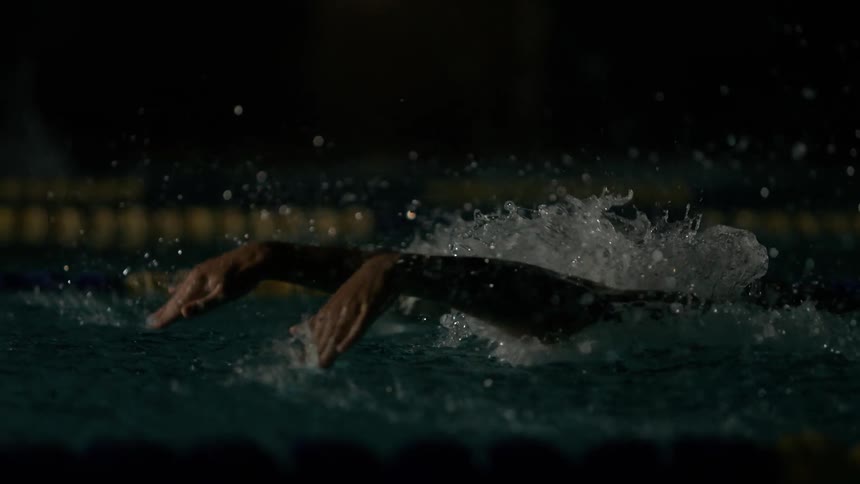 A person wearing a swimming cap and goggles is swimming in a pool and his arm goes into the water and lifts a lot of water forward