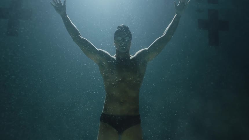 Man wearing a swimming cap and goggles at the bottom of a pool opening his arms and rising towards the surface