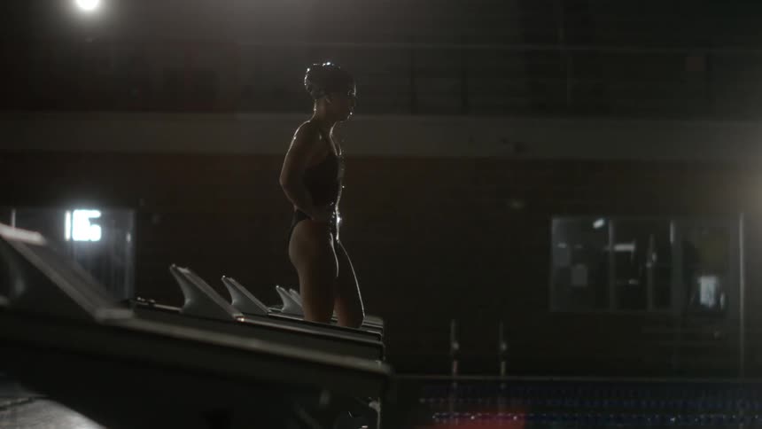 A woman is standing in front of a swimming pool and she is wearing swimming clothes and has water dripping off her