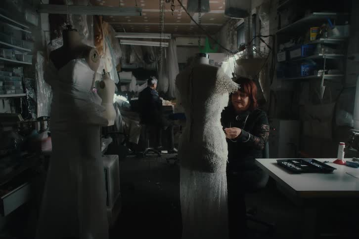 A seamstress is fitting dresses over a pair of mannequins in a cluttered dimly lit workshop