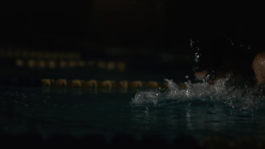 A male wearing a cap and goggles is swimming in darkness creating large splashes