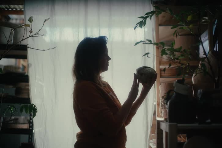 A silhouette of a woman stands in front of a window holding a small decorative object in a dimly lit room