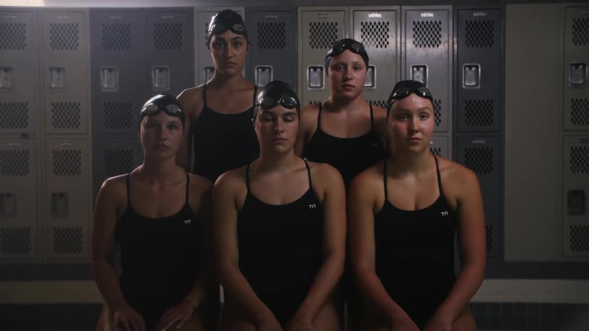A group of young women dressed in black swimwear sit closely together in front of a row of grey lockers