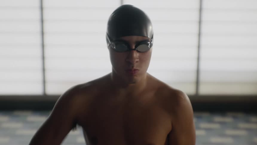 A shirtless man with a swimming cap and goggles is stretching his arms