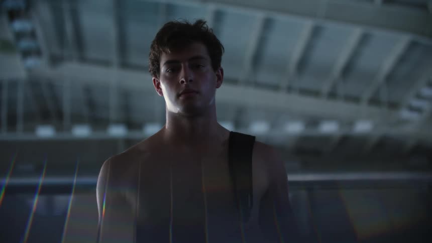 A young man in a gym not wearing a shirt carrying a bag by a strap on his shoulder looking sad