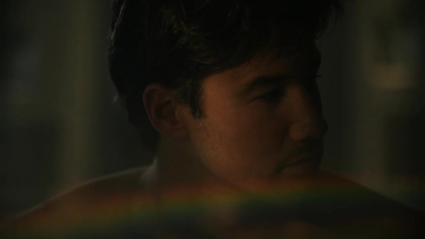 A man wearing no shirt looks to the side as lights create a rainbow on his chest