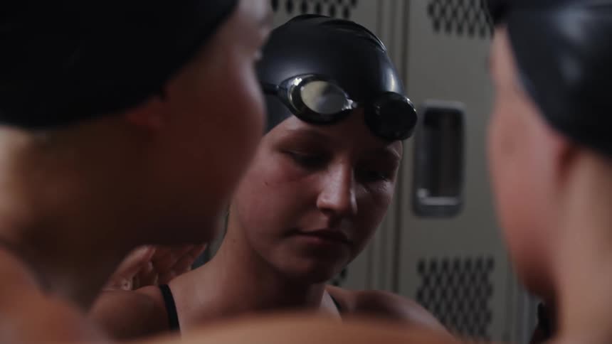 Three young women in a locker room wearing swimming gear with two of them cheering up a third