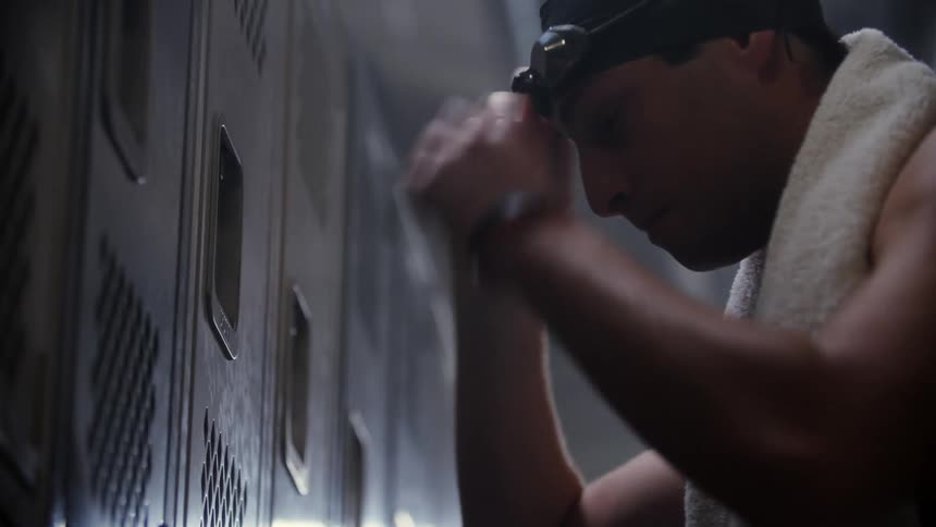 A shirtless man with a towel over his shoulder and a swimming cap is tired and slaps a locker and leaves