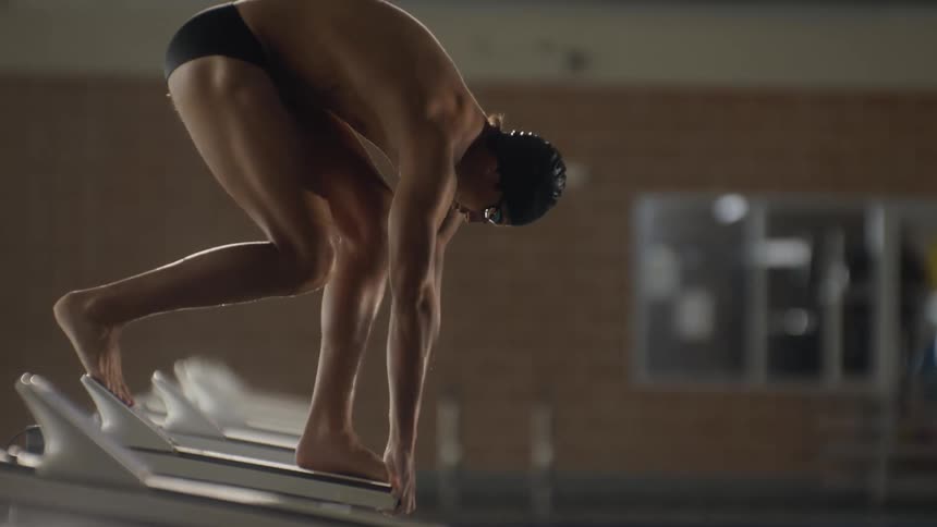 A swimmer poised on a starting block at the edge of a pool jumps in and dive