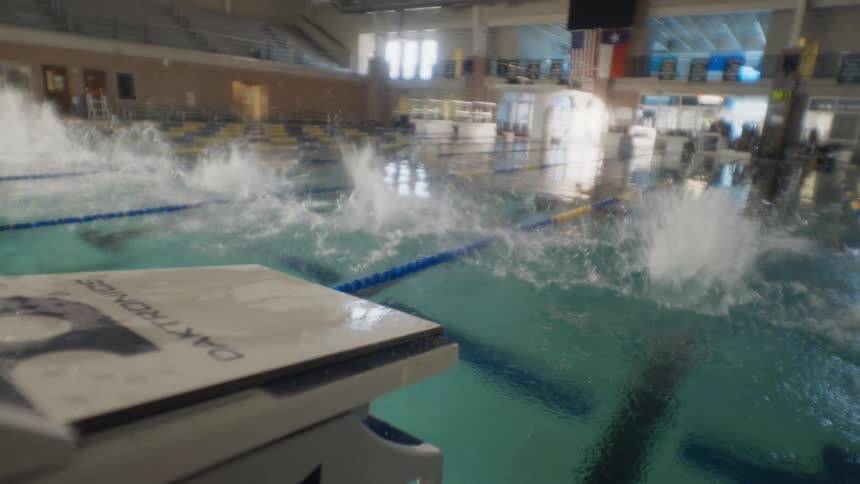 Four competitive male swimmers dive into an indoor pool at the same time