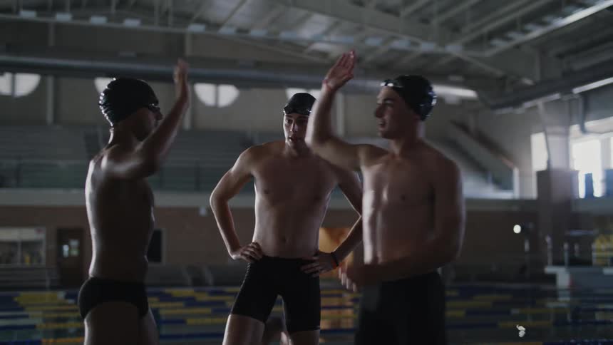 Three swimmers stand on a swimming platform in an indoor pool giving each other high fives