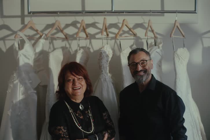 A man and woman pose in front of wedding dresses in the shop