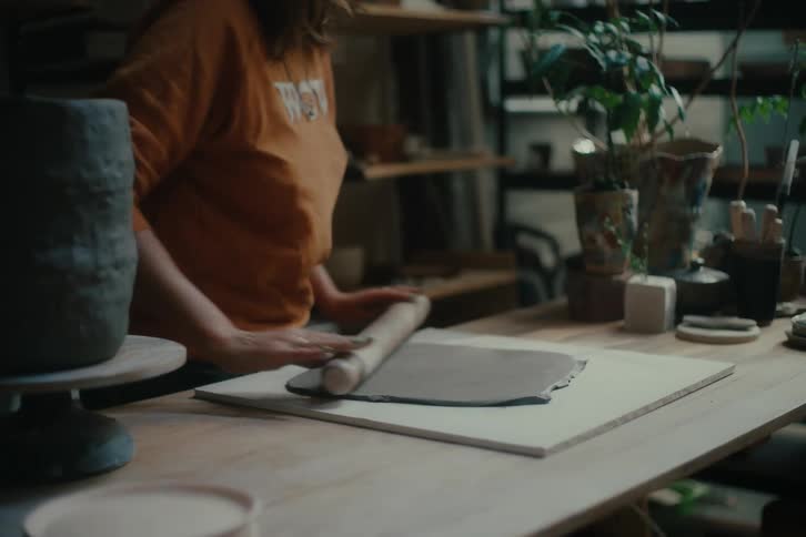 Woman working with clay at a table surrounded by pottery tools and plants