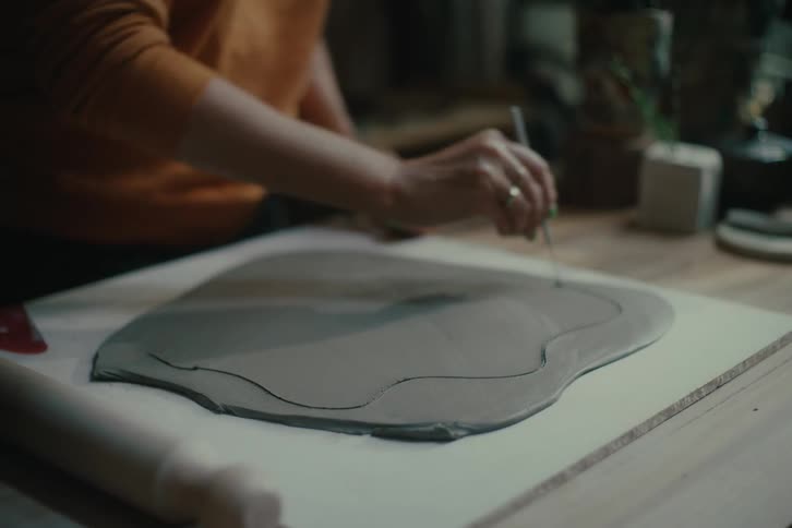 A persons hands work on a piece of clay atop a table cutting a shape with a tool