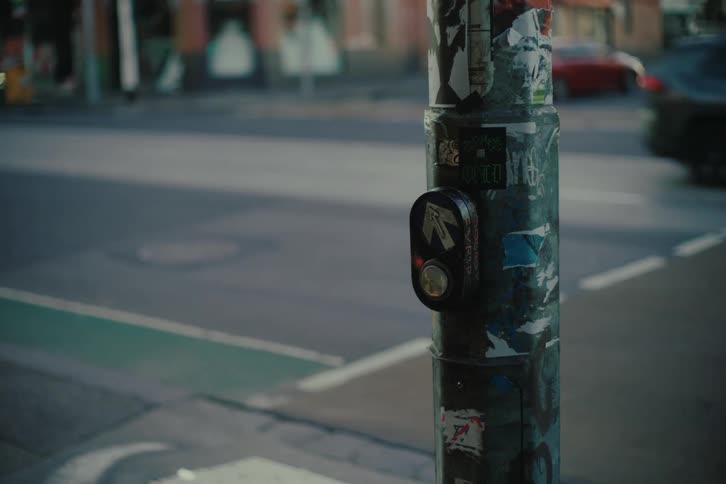 Pedestrian signal button covered with stickers on an urban pole with vehicles passing by in the background