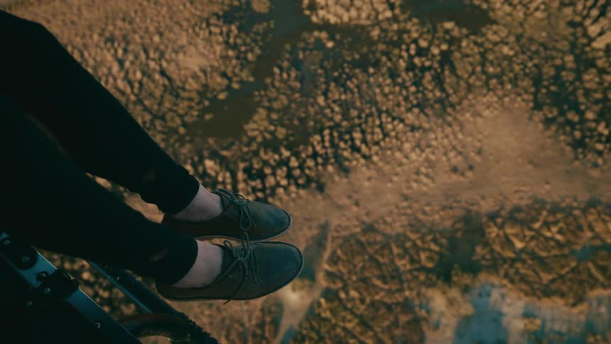 A person dangles their feet out of a helicopter flying over a forest with bodies of water and open grassy plains