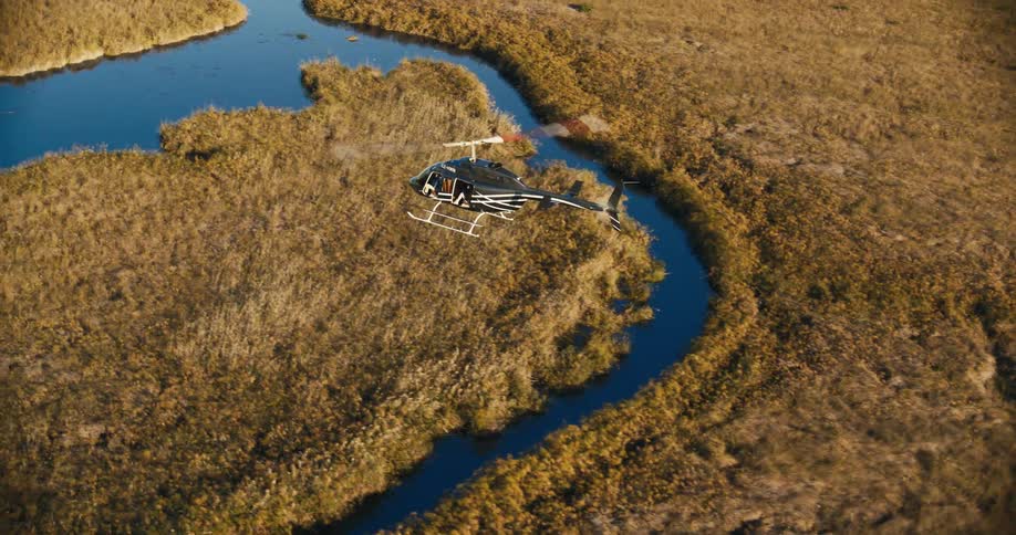 A black helicopter flying over a savannah with some stretches of a river