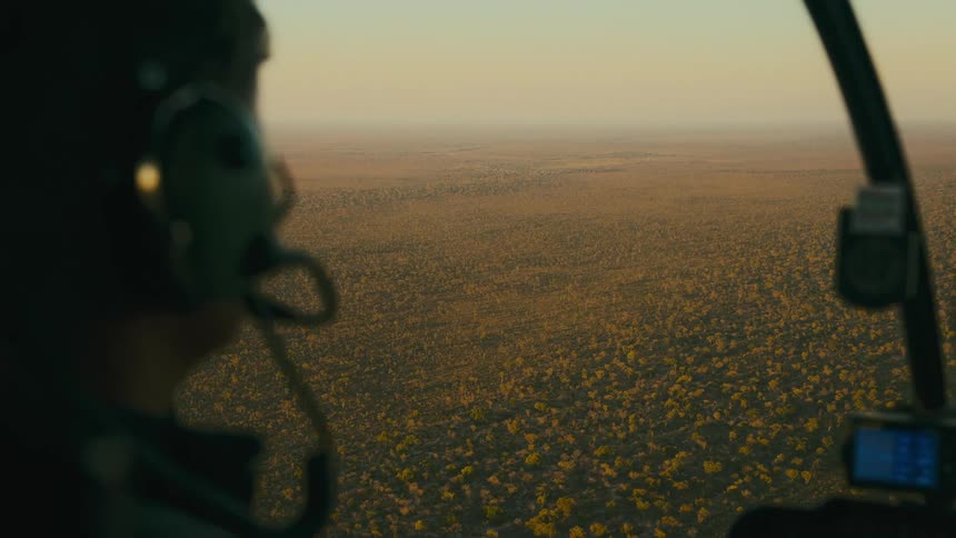 A male pilot steers a helicopter over a vast savanna