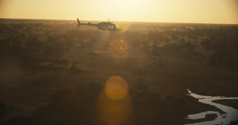 A helicopter flying over a savannah with a river and the sun is shining brightly in the sky