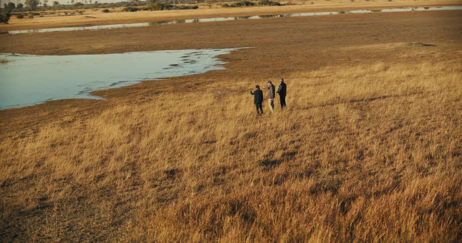Three people walk through a vast area of dry grassland with patches of water in the background