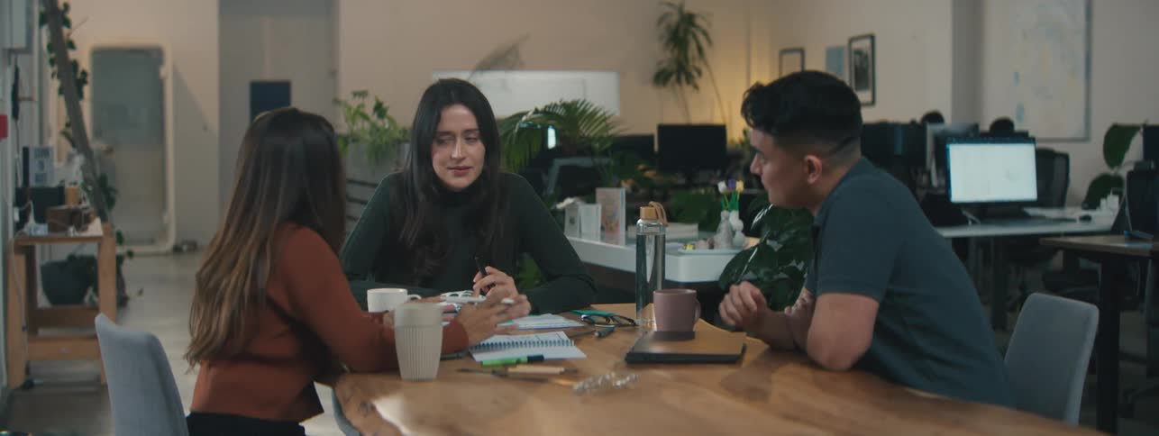 Three people are sitting around a table in an office environment engaged in a discussion