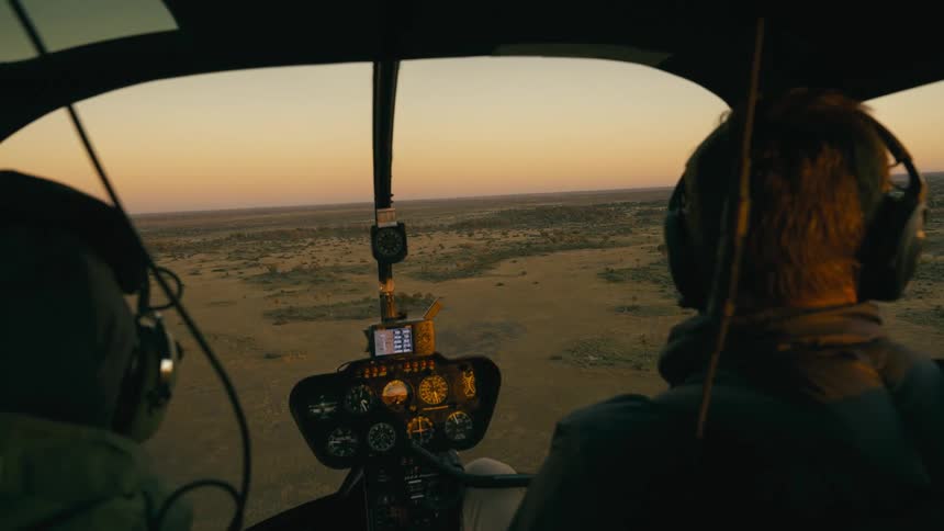 The backs of two pilots flying a helicopter in a natural region
