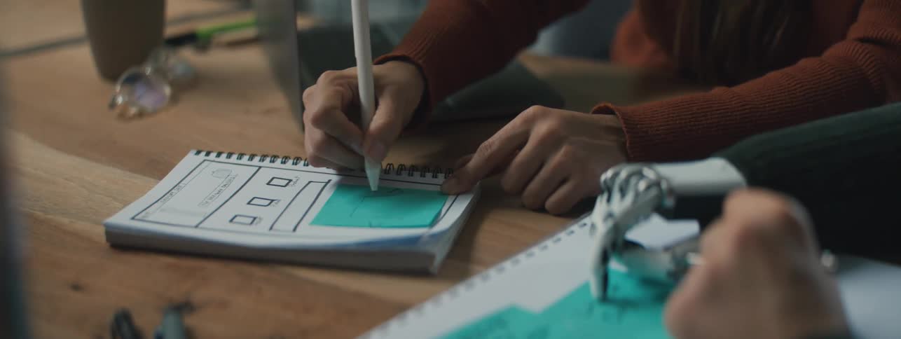 A persons hand writing something on a blue postit stuck on a notebook and a person with a robotic hand on the right side