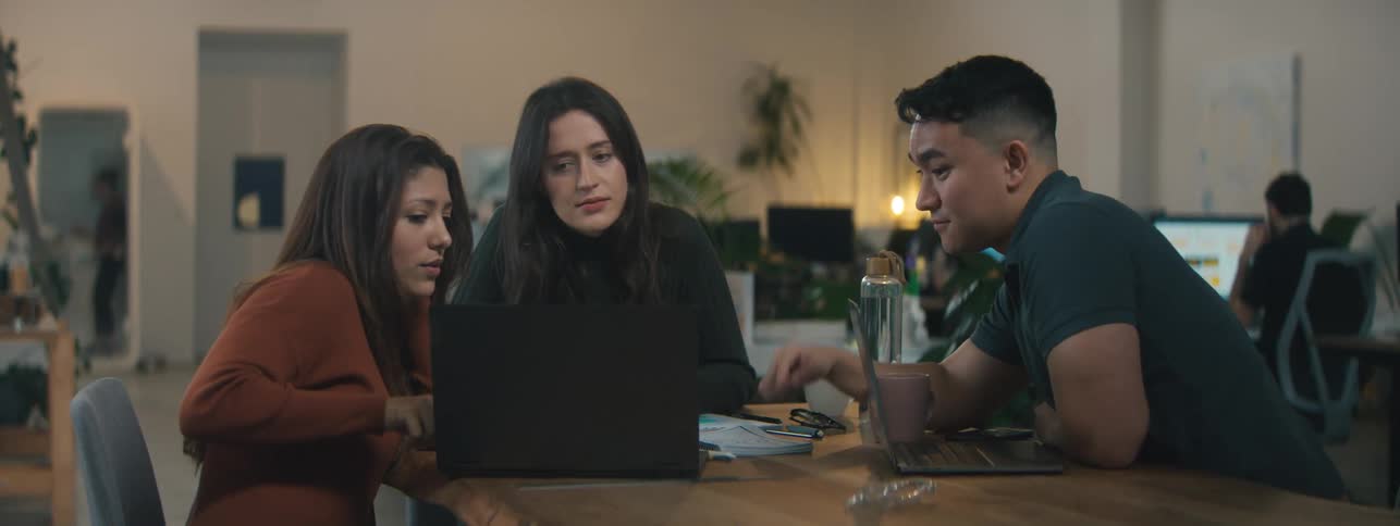 Two women and a man are sitting around a table and looking at a laptop while talking
