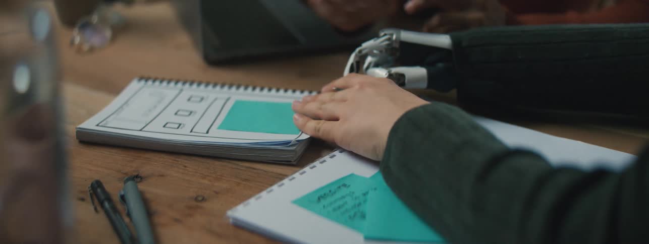 Hands of two women with one of them robotic writing in a notebook