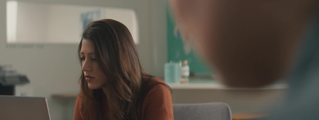 The face of a young woman who is looking straight ahead where there is a laptop on the table and she moves closer to the screen