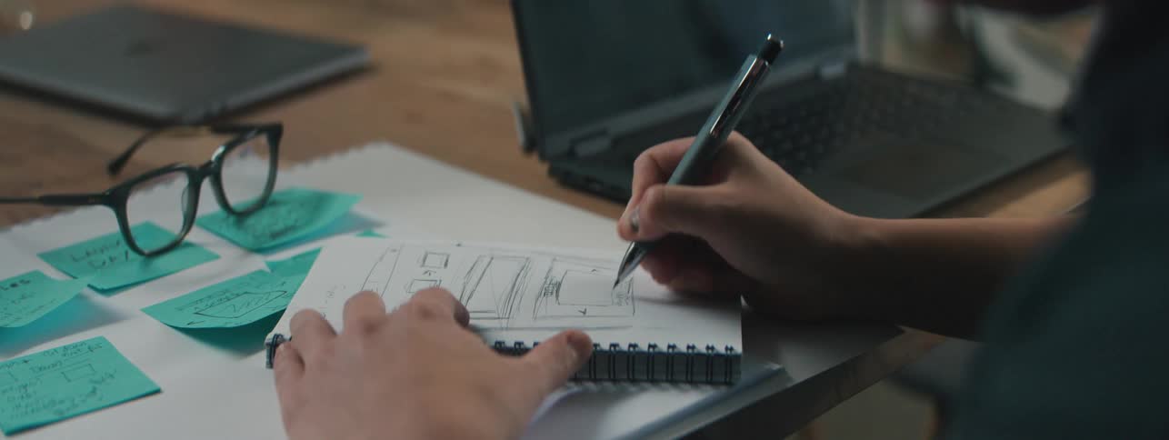 A person sketching in a notebook surrounded by sticky notes and a laptop