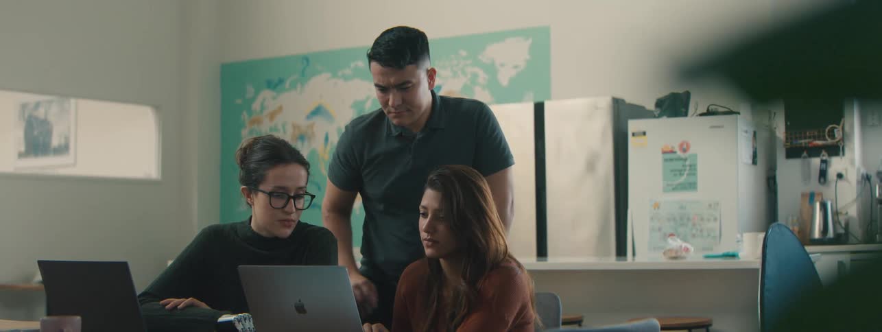 Two women sitting at a table with laptop and a colleague standing interacting