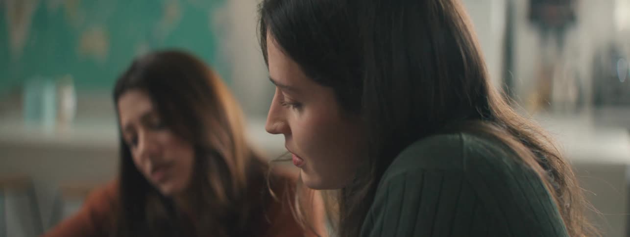 Two women with long brown hair sit at a table while talking with someone else