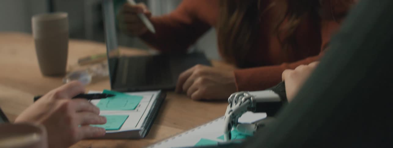 A human hand writing something on a paper and a woman starts pointing to the paper and talking