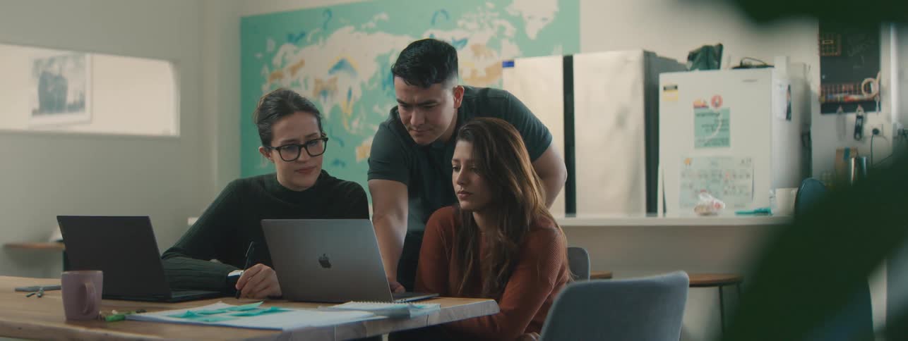 Three people collaborating at a table with laptops focused on a computer screen in a bright modern workspace