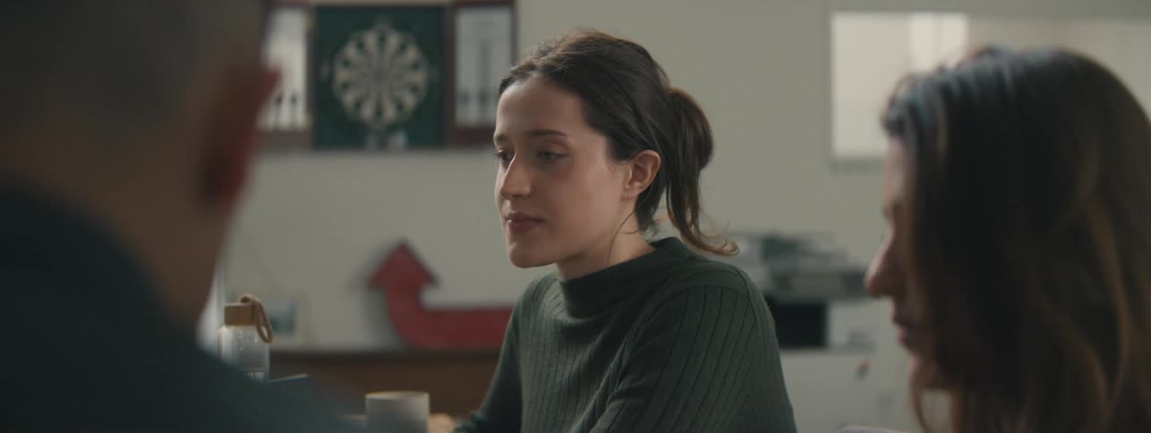 A young woman in a green coat is sitting next to some people and she takes off her reading glasses and looks thoughtful