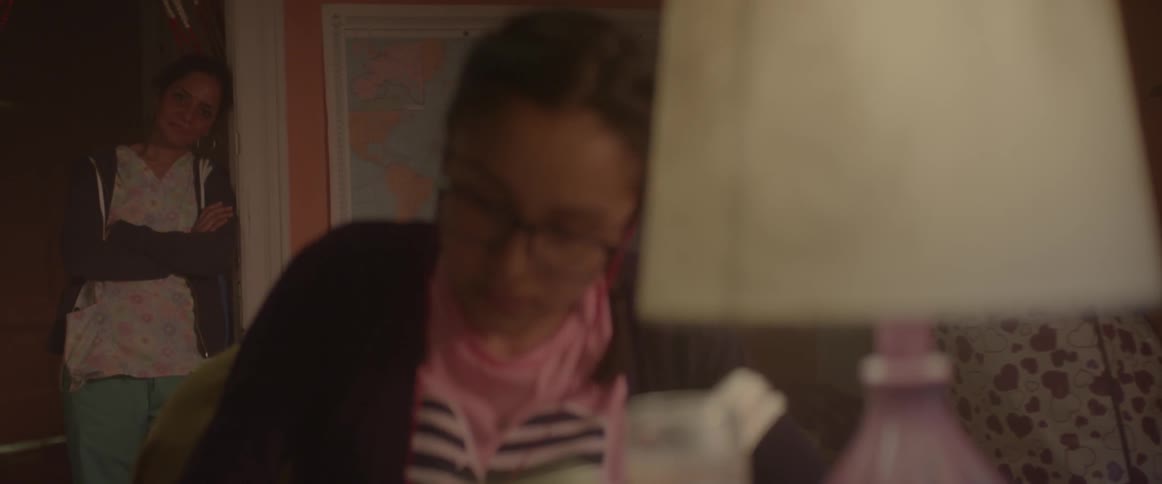 Spectacled woman writing at a table  with a person watching from behind in the doorway