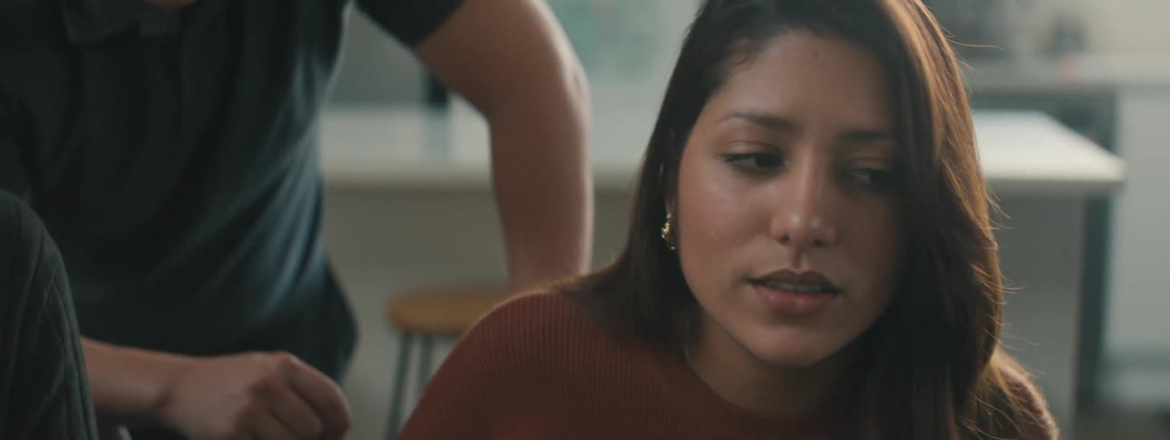 A young woman sitting while explaining something to someone sat next to her and a man standing behind her