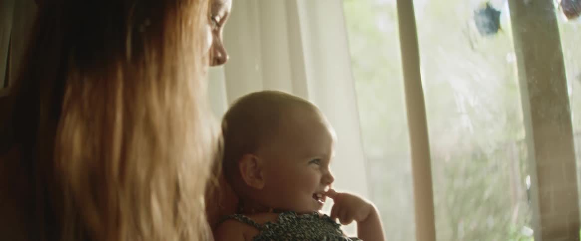 A baby blushes and laughs at her daddy off screen whilst her mother holds her happily