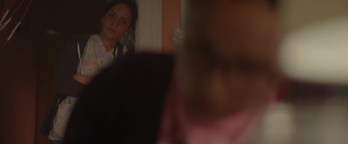 Spectacled woman writing at a table  with a person watching from behind in the doorway