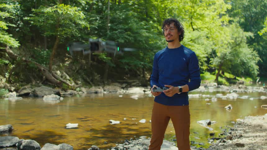 An individual stands by a creek holding a remote control flying a drone