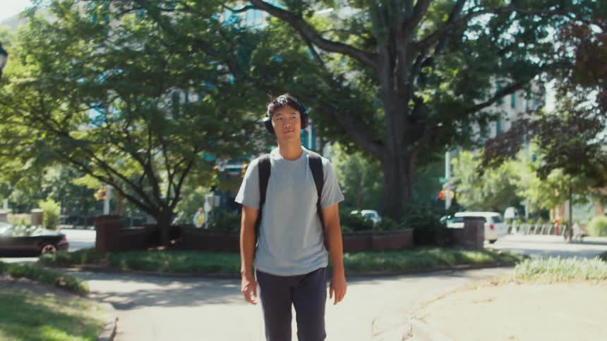 A young asian man walks down a city sidewalk while wearing headphones